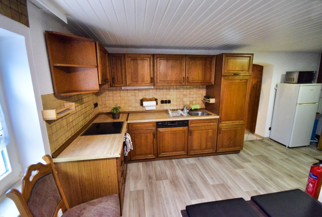 Sie sehen eine gewinkelte Küche in Naturholz mit Cerankochfeld, Spüle sowie Kühlschrank, rechts in der Ecke steht eine Kühl-Gefrierkombination.