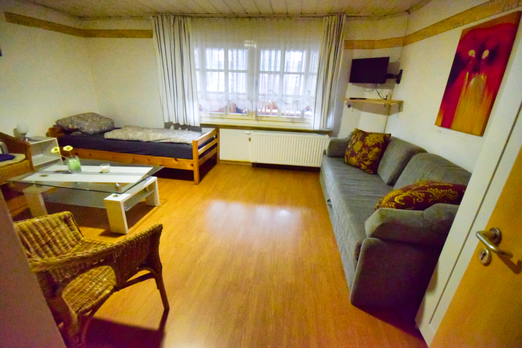Sie sehen zwei Einzelbetten in einem Monteurzimmer, die Betten sind aus Kieferholz, sauber und gepflegt. Eine blaue Couch an der rechten Wand und ein Tisch in der Mitte.