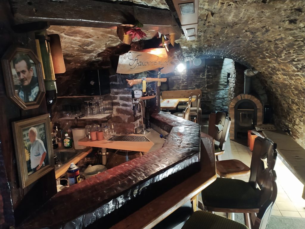 Gewölbekeller mit Theke und im Hintergrund ein Ofen davor Sitzgelegenheiten.